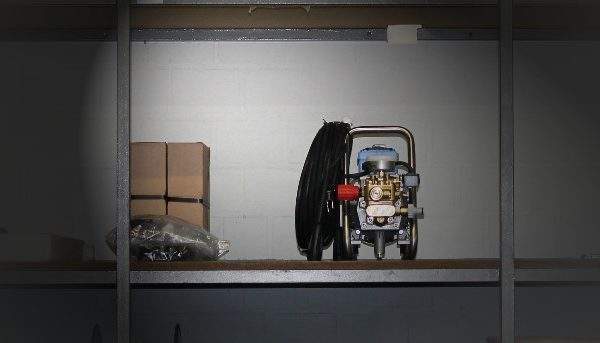 Hochdruckreiniger auf einem Regal in einem Lagerraum, umgeben von Kartons und einem Schlauch.